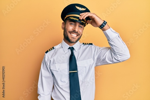 Handsome hispanic man wearing airplane pilot uniform smiling confident touching hair with hand up gesture, posing attractive and fashionable