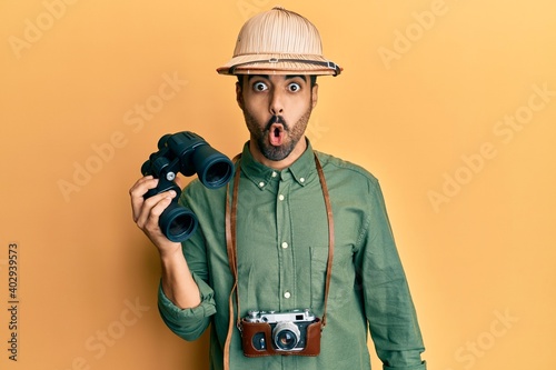 Young hispanic man wearing explorer hat looking through binoculars scared and amazed with open mouth for surprise, disbelief face