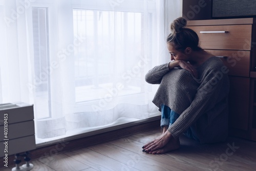 A devastated sad woman is sitting on the floor in her living room looking at the window. She is depressed.