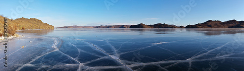 Frozen Baikal Lake on a sunny winter day. Panoramic view of beautiful blue smooth ice with cracks in Kurkut Bay. Tourists walking and skating on ice are visible in the distance. Natural background