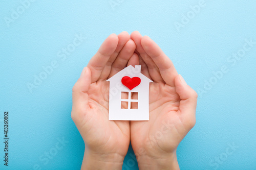 White paper house with bright red heart in young adult woman palms on light blue table background. Pastel color. Closeup. Point of view shot. Top down view.