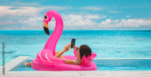 Relaxing woman floating in flamingo inflatable swimming pool toy at luxury resort using mobile phone sunbathing. Caribbean travel vacation hotel lifestyle.