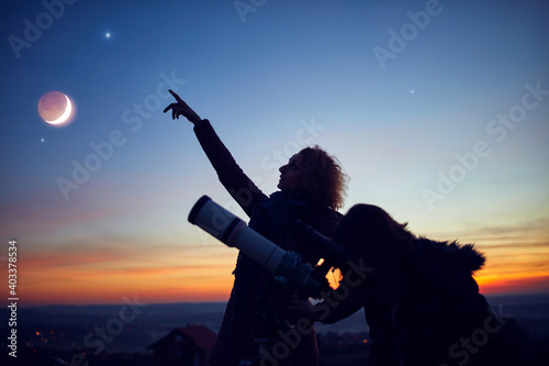 Mother and daughter observing stars, planets, Moon and night sky with astronomical telescope.