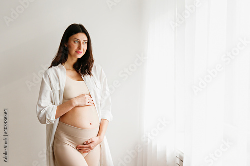 Portrait shot of young beautiful woman on second trimester of pregnancy. Close up of pregnant female in casual attire with arms on her round belly. Expecting a child concept. Background, copy space.