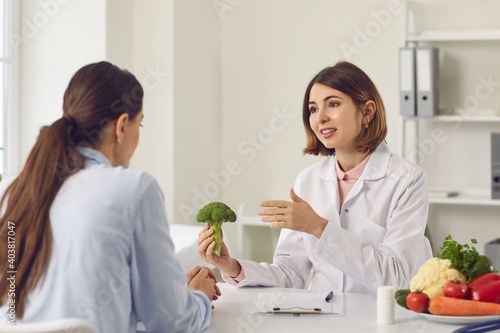Qualified female dietitian, nutritionist, healthy food expert, doctor of alternative medicine, consultant in health center holding broccoli and telling young woman about benefits of eating vegetables
