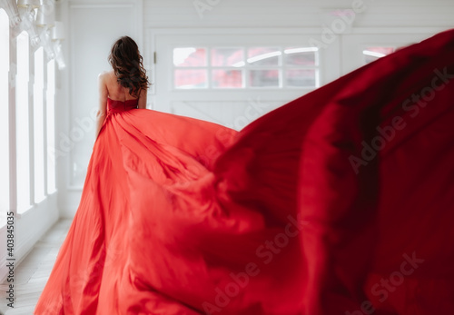 Brunette woman with long hair standing by her back in long red evening dress in white room.