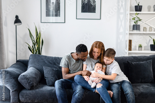 Friendly multiracial of four family spends time together in cozy living room. A mother, a father, a school-age son and baby girl sit in embraces on the comfortable sofa and laugh