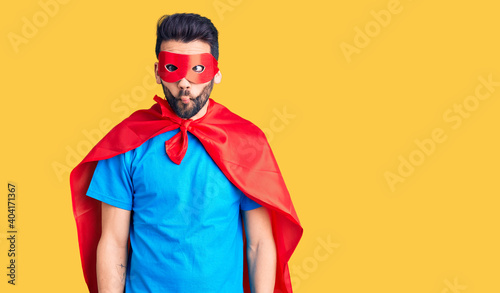Young handsome man with beard wearing super hero costume making fish face with lips, crazy and comical gesture. funny expression.