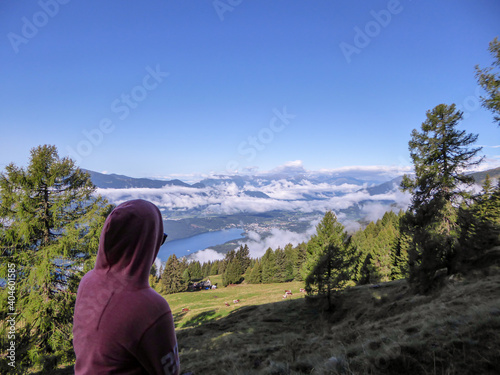A woman in an orange hoodie enjoying the idyllic view on Alpine Millstaettersee lake in Austria. She is hiking in high Alps. The valley is shrouded with some clouds, sunny day. The girl is calm, happy