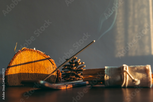 Burning incense stick in a scene natural and wooden decoration objects during sunset. Selective focus on stick tip ash. Cozy home environment.