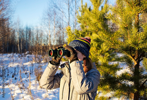 Young woman birdwatcher in winter clothes and knitted scarf looking through binoculars in winter snowy pine forest. Ecology and ornithological research, birdwatching
