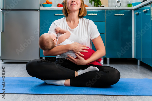 A young smiling mother meditates in the kitchen, sitting on a sports mat, holding the baby in her arms. Close up. The concept of yoga and meditation