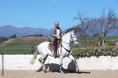 Young guy in casual outfit riding white horse on sandy ground