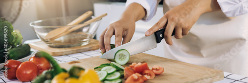 Woman using knife and hands cutting cucumber on wooden board in kitchen room.