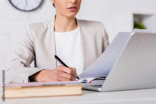 cropped view of translator writing in notebook while holding document near laptop, blurred foreground
