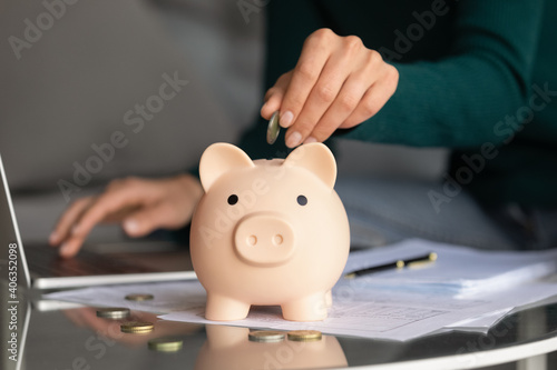 Close up of woman put drop coin into piggy bank managing budget paying bills taxes online on computer. Female feel provident economical take care of expenses, save money or make investment.