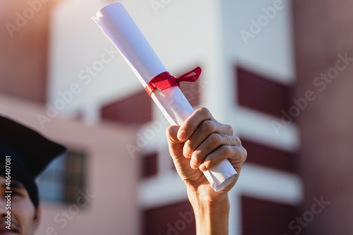 Close-up shot of a university graduate holding degree certification to shows and celebrate his success in the college commencement day with sunlight in the background.