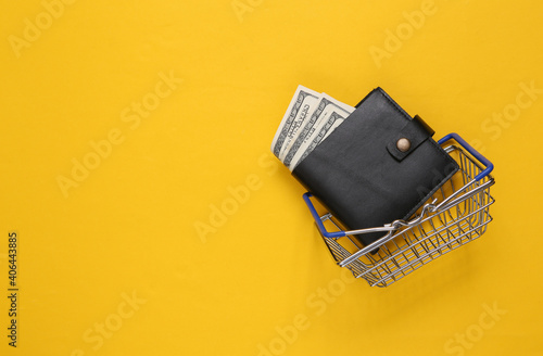 Leather wallet with one hundred dollar bills and shopping basket on yellow background. Shopping concept