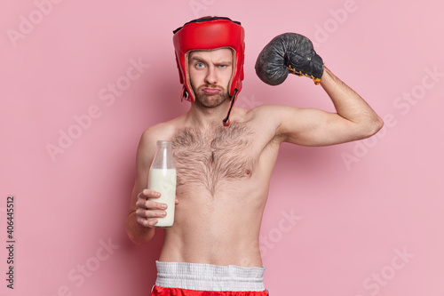 Serious man boxer with skinny body wears boxing gloves and hat raises arm shows muscles drinks milk for having strong biceps demonstrates his strength and power. Sport and motivation concept