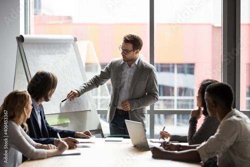 Skilled 30s caucasian leader in eyeglasses coaching interested business people, giving educational workshop presentation in office. Concentrated employees listening to professional coach at workshop.