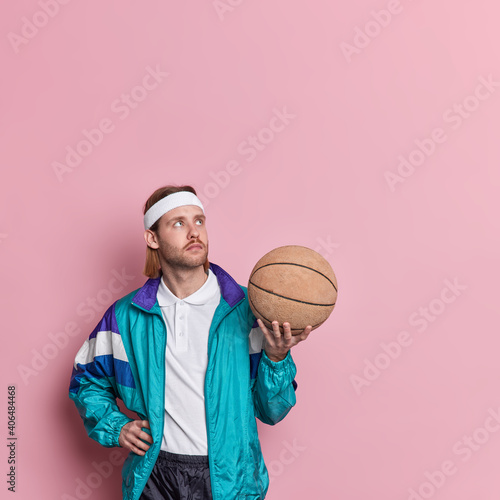 Thoughtful professional basketball player in activewear holds ball concentrated above going to play favorite game isolated over pink background. Hobby leisure sport concept. Active guy indoor