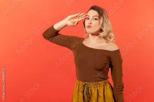 Portrait of responsible serious woman with blonde hair in brown blouse saluting commander, listening order with obedient expression. Indoor studio shot isolated on red background