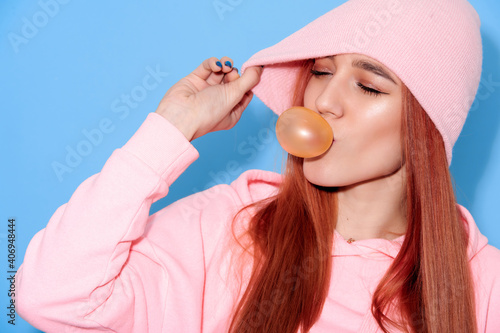 Young attractive girl in pink clothes blowing bubbles from chewing gum on blue background