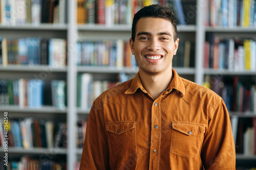 Portrait of successful hispanic business man looks directly at the camera, smile. Happy male freelancer or student standing in modern office or university library