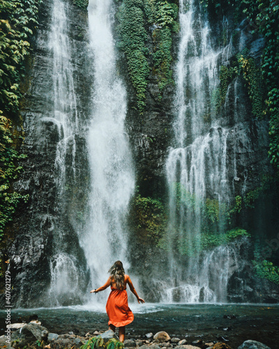 Young woman standing in front of a very big and beautiful waterfall. Dancing next to sekumpul waterfall in bali, indonesia. 