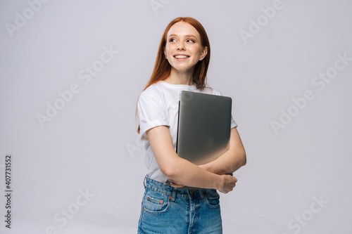 Smiling young woman student holding laptop computer and looking away on isolated gray background. Pretty lady model with red hair emotionally showing facial expressions in studio, copy space.