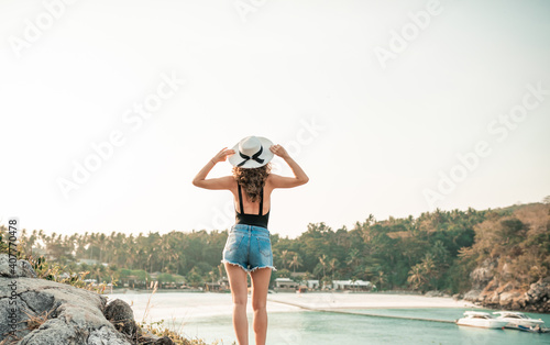 Dreamy woman in sexy black swimsui tsitting on a big stone at tropical beach and wicker hat. Summer vacation travel. Lifestyle concept