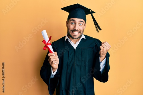 Young hispanic man wearing graduation robe holding diploma screaming proud, celebrating victory and success very excited with raised arm