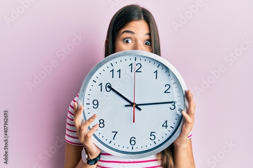 Young brunette woman holding big clock covering face celebrating crazy and amazed for success with open eyes screaming excited.