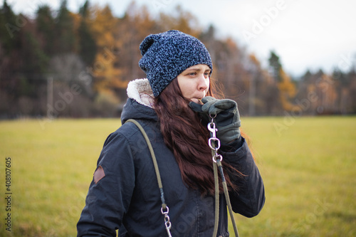 young dog breeder walk her dog. Detail on attention of a brunette who whistles disobedient dog. Woman aged 20-24 holds whistle and watches bitch. Candid portrait in authentic setting in grey moss tone