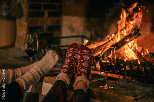 Cosy winter evening. Drinking wine and reading book next to fireplace. Feets in woollen socks by the Christmas fireplace