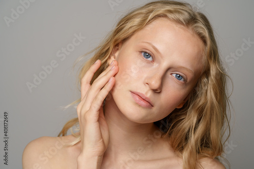 Closeup of pretty young woman face with blue eyes, curly natural blonde hair, has no makeup, touching her soft skin, standing shirtless with bare shoulders, looking at camera. Studio grey background