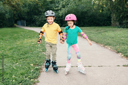 Happy Caucasian friends boy and girl in helmets riding on roller skates in park on summer day. Support and help from friend. Seasonal outdoors children activity sport. Kids individual summer sport.