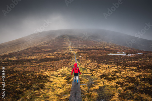 Boy in a red jacket, hiking on wooden path leading through the wicklow mountains, Djouce pek Ireland. Wooden path in foggy mountain landscape, in Autumn. 2019 Ireland