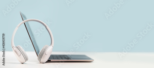 Side view of slim laptop with wireless headphones on grey desk. Blue background. Distant learning. working from home, online courses or support. Audio podcast. Helpdesk or call center headset