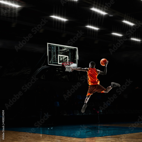 Champion. African-american young basketball player in action and motion in flashlights over dark gym background. Concept of sport, movement, energy and dynamic, healthy lifestyle. Arena's drawned.