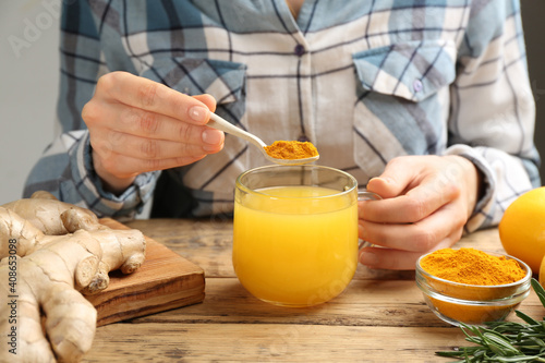 Woman adding turmeric to immunity boosting drink at wooden table with ingredients, closeup