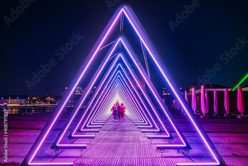 Purple coloured gate of light or purple light tunnel installation made of triangular neon and led lights at night as blurred people walk through the bright deep passage that resembles a human trachea