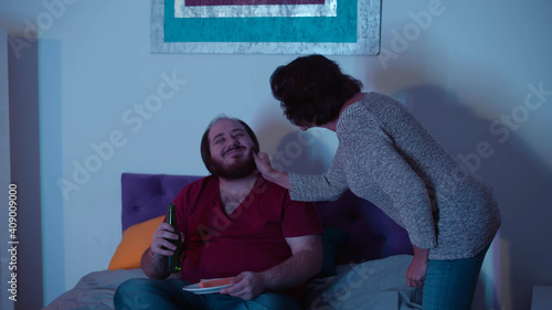 Young fat guy watching tv in bed while senior mother bringing him beer and sandwich