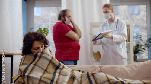 Sick aged woman covered in blanket lying on couch while adult son in safety mask consulting doctor