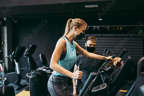 Young fit and attractive woman at body workout in modern gym together with her personal fitness instructor or coach. They keeping distance and wearing protective face masks. Coronavirus sport theme.
