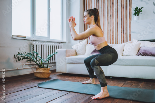 Young fit woman in sportswear do squat exercise indoors in living room. Online indoors workout. Training at home