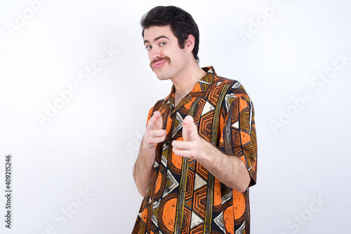 Joyful Young caucasian man wearing generic pattern printed shirt against yellow wall wink and points index fingers at camera, chooses someone, makes finger gun pistol.