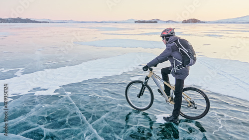 Man is riding bicycle on the ice. Ice of the frozen Lake Baikal. Teenage is dressed in black down jacket, cycling backpack, helmet. Tires on covered with special spikes. Traveler boy is ride cycle.