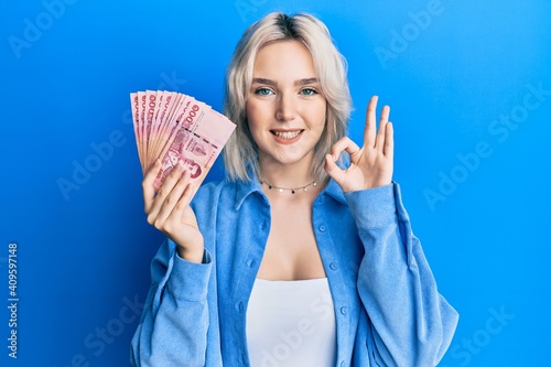 Young blonde girl holding thai baht banknotes doing ok sign with fingers, smiling friendly gesturing excellent symbol