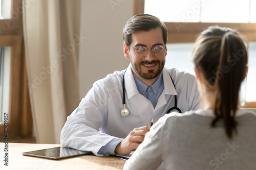 Young Caucasian male doctor in white uniform talk consult female patient in private hospital or clinic. Smiling man GP have consultation with woman, discuss anamnesis or results. Healthcare concept.